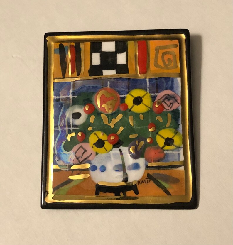 May include: A square ceramic tile with a black frame and gold trim. The tile features a colorful painting of a vase with flowers in shades of yellow, red, and blue. The artist's signature, "Tomz", is visible in the lower right corner.