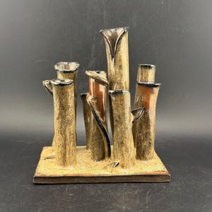 May include: A ceramic sculpture with multiple vertical, cylindrical shapes in brown, tan, and black. Some cylinders have a reddish-brown accent. The sculpture sits on a rectangular base with a textured surface, creating a unique decorative piece.