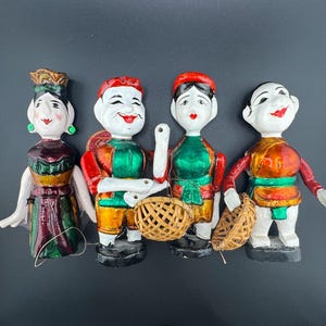 May include: Four traditional Vietnamese water puppet figures. The figures are made of painted wood and have colorful clothing. They are holding baskets and have exaggerated facial features.