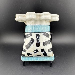 Può includere: Un vaso in ceramica bianca con un design astratto blu e nero. Il vaso ha un'ampia parte superiore a forma di conchiglia e una base quadrata. È su un supporto in metallo nero.