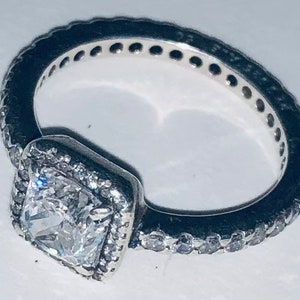 May include: A silver ring with a square-shaped clear stone set in a silver frame. The ring is adorned with small clear stones around the frame and the band.