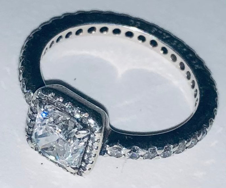 May include: A silver ring with a square-shaped clear stone set in a silver frame. The ring is adorned with small clear stones around the frame and the band.