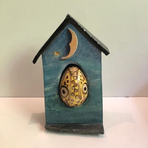 Può includere: Una casa in ceramica con un esterno dipinto di blu e bianco. La casa ha una luna crescente sul davanti e un uovo di ceramica giallo con un viso all'interno della finestra.