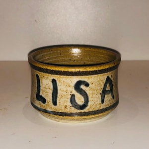 以下が含まれることがあります： 濃い茶色の縁取りと側面に青い文字で「LISA」と書かれた、小さな茶色の陶器のボウルです。