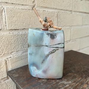 May include: A ceramic vase with a light blue, green, and brown glaze. The vase has a unique, organic shape and is topped with a small piece of driftwood.