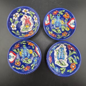 Puede incluir: Juego de cuatro cuencos pequeños de cerámica azul con diseños florales coloridos. Cada cuenco presenta un tulipán blanco con hojas verdes y detalles rojos, amarillos y azules. Los cuencos son perfectos para servir aperitivos, salsas o condimentos.