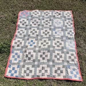 May include: A handmade quilt with a repeating pattern of gray and white squares, accented with blue squares. The quilt has a red border and is laid out on a grassy surface. The quilt is a vintage item.