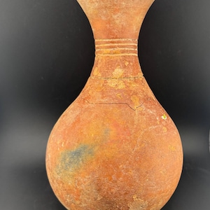 May include: A terracotta-colored vase with a bulbous body and a flared rim. The vase has a textured surface with horizontal ridges near the neck. The overall shape is reminiscent of ancient pottery, with visible cracks and imperfections.