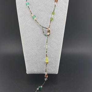 May include: A long beaded necklace with a Y-shaped pendant. The necklace features a variety of colorful beads, including clear, green, brown, and orange. The beads are strung on a dark cord and displayed on a gray mannequin.