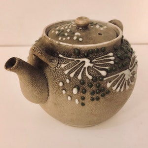 Może przedstawiać: Mały, szary ceramiczny czajnik z białym i zielonym wzorem kwiatowym. Czajnik ma okrągły korpus i mały dzióbek.
