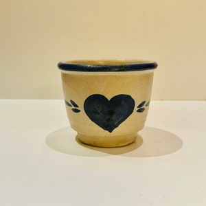 Puede incluir: Un pequeño cuenco para huevos de cerámica de color crema con un diseño de corazón negro y un borde azul.