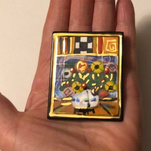 May include: A colorful ceramic tile with a gold frame depicting a still life of flowers in a vase. The tile has a black and white checkered pattern at the top and a gold swirl design in the upper right corner.