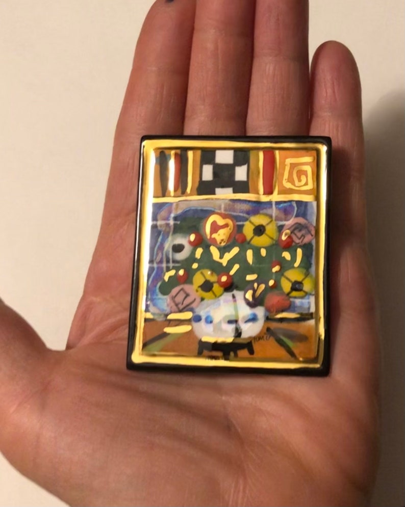 May include: A colorful ceramic tile with a gold frame depicting a still life of flowers in a vase. The tile has a black and white checkered pattern at the top and a gold swirl design in the upper right corner.