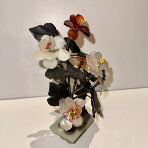 May include: A decorative floral sculpture featuring white, brown, and translucent flowers with yellow and red accents. The flowers are set on dark green leaves and stems, mounted on a rectangular stone base. The sculpture casts a shadow.