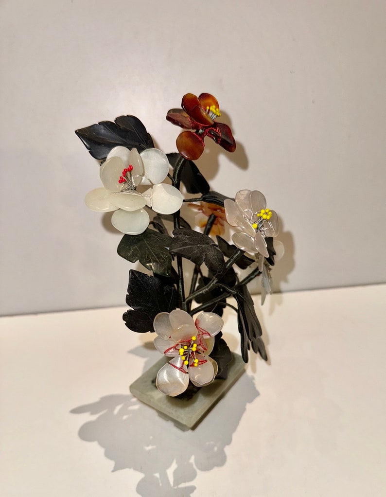 May include: A decorative floral sculpture featuring white, brown, and translucent flowers with yellow and red accents. The flowers are set on dark green leaves and stems, mounted on a rectangular stone base. The sculpture casts a shadow.