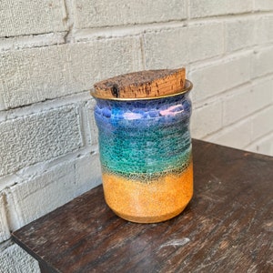 Könnte beinhalten: Eine Keramikdose mit einem Korkdeckel. Die Dose hat eine blaue, grüne und gelbe Glasur mit einer craquelé-Oberfläche. Die Dose steht auf einer Holzoberfläche.