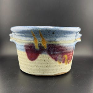 May include: A handmade ceramic bowl with two handles. The bowl is glazed in shades of blue, white, and purple with accents of yellow.