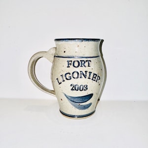 Può includere: Una tazza in ceramica bianca con macchie blu e un design blu. La tazza ha un manico e dice "FORT LIGONIER 2003" sul davanti.