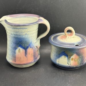 May include: A ceramic creamer pitcher and sugar bowl set. The pitcher has a handle and spout, and the sugar bowl has a lid with a small handle. Both pieces are painted with blue, pink, and white abstract designs.
