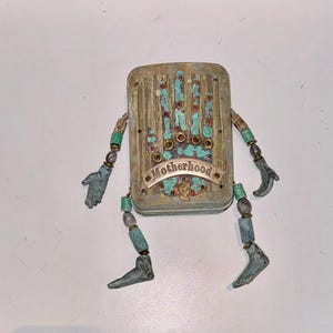 Könnte beinhalten: Eine kleine, rustikale Metallfigur mit handbemaltem Design. Die Figur hat einen rechteckigen Körper mit einem türkisfarbenen und goldenen Design. Die Figur hat zwei Arme und Beine aus Perlen und Metall. Die Figur trägt den Schriftzug "Motherhood" auf einem Banner.
