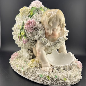 Puede incluir: Figura de porcelana de un querubín rodeado de flores. El querubín está arrodillado, sosteniendo un plato en forma de concha. Las flores son en tonos rosa, amarillo y blanco, con hojas verdes. La base es ovalada.