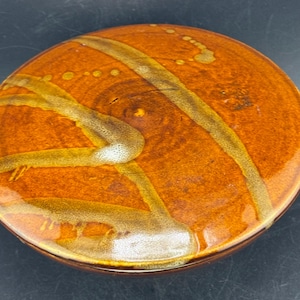 May include: A round ceramic lid with a glossy finish in shades of burnt orange and deep brown. The surface features abstract, sweeping brushstroke patterns in a lighter tan hue, along with small, scattered circular drip-like markings.