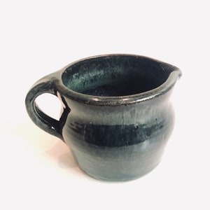 May include: A small, dark green ceramic creamer with a rounded body and a handle. The creamer has a glossy finish.