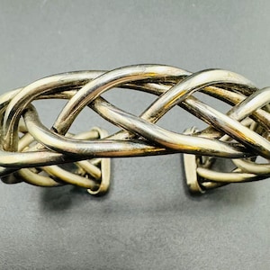 Puede incluir: Un brazalete de plata trenzado con un diseño tejido. El brazalete está hecho de alambre de plata grueso y retorcido.