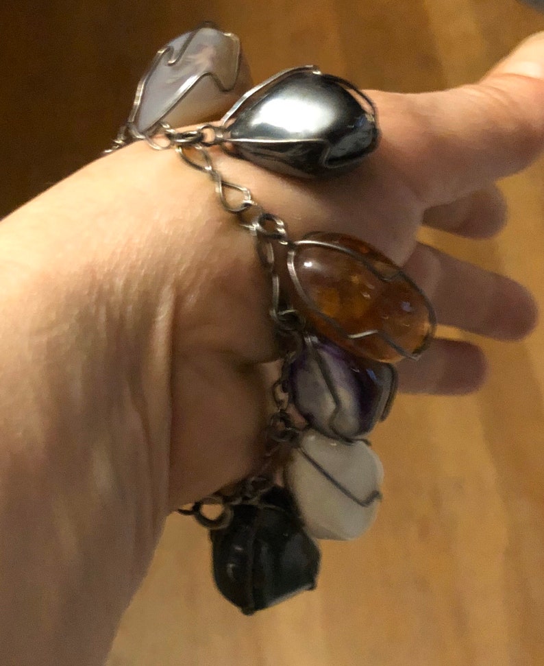 Pode incluir: Um bracelete de corrente de prata com v&aacute;rios pingentes em forma de cora&ccedil;&atilde;o de pedras preciosas. Os pingentes s&atilde;o de v&aacute;rias cores, incluindo branco, cinza, &acirc;mbar e roxo.
