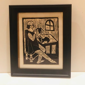 Op de afbeelding: Zwart-witte houtsnede van een persoon die aan een tafel zit met een klein figuurtje op schoot. De persoon draagt een lange jurk en een hoed op zijn hoofd. De print is ingelijst in een zwart frame.