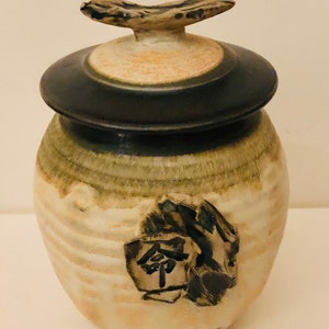 May include: A ceramic jar with a lid, featuring a brown and white glaze with a black Japanese character on the front. The jar has a unique, irregular shape and a textured surface.