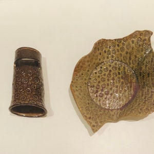 May include: Two ceramic dishes with a brown glaze and a swirl pattern. The larger dish is a shallow bowl with a wavy edge. The smaller dish is a cylinder with a small opening on the side.