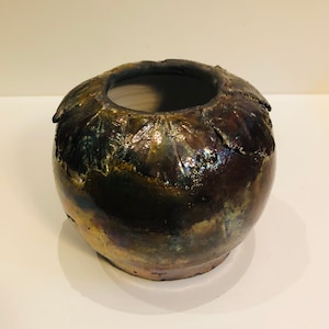 Puede incluir: Un pequeño jarrón redondo de cerámica con un esmalte metálico texturizado en tonos marrones, dorados y negros. El jarrón tiene una abertura ancha y un borde ligeramente acampanado.