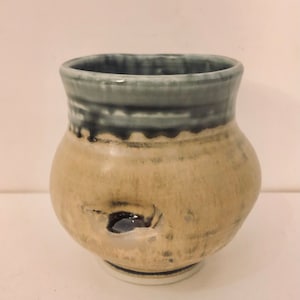 May include: A small, round, ceramic vase with a light brown body and a blue and black glaze around the rim. The vase has a small hole in the side.