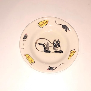 Puede incluir: Un plato de cerámica blanco con un diseño pintado a mano de un gato siamés, tres ratones y cuatro trozos de queso. El gato está tumbado y tiene los ojos azules.