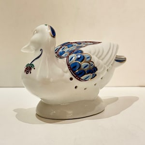 Könnte beinhalten: Eine weiße Keramik-Entenfigur mit blauen, goldenen und burgunderfarbenen Akzenten. Die Ente hat detaillierte Flügelmuster und dekorative florale Designs. Die Figur hat kleine Löcher und steht auf einem abgerundeten Sockel.