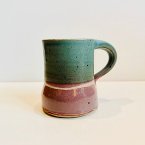 May include: A handmade ceramic mug with a teal upper section and a deep purple lower section. A thin white and pink band separates the two colors. The mug has a curved handle and is suitable for hot or cold beverages.