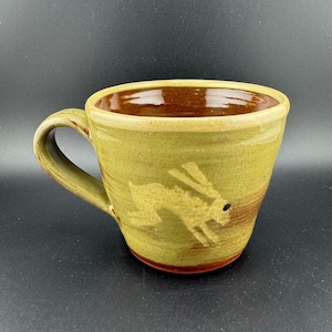 Op de afbeelding: Een groene keramische mok met een bruine rand en een wit konijn ontwerp. Het konijn kijkt naar links en heeft een zwart oog.