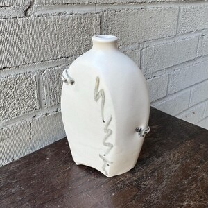 Könnte beinhalten: Weiße Keramikvase mit einer einzigartigen, asymmetrischen Form und einem dezenten, abstrakten Design in Grau. Die Vase hat eine schmale Öffnung und einen breiten Boden.