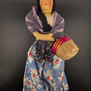 Può includere: Bambola fatta a mano con volto dipinto, capelli neri e uno scialle viola. La bambola indossa un vestito blu con stampa floreale e porta un piccolo cestino intrecciato con accenti rosa e gialli. È un'opera d'arte popolare.