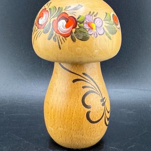 Könnte beinhalten: Dekoratives Objekt in Pilzform aus Holz. Die Oberseite ist mit einem Blumendesign mit roten, weißen und lila Blumen verziert. Der Stiel hat ein schwarzes, stilisiertes Blattdesign. Der Pilz ist aus hellem Holz gefertigt.