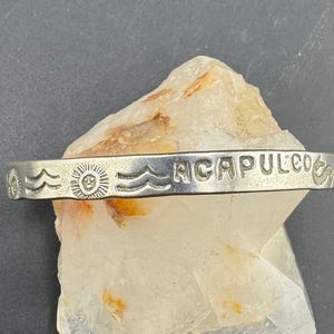Op de afbeelding: Een zilverkleurige armband met het woord "ACAPULCO" in hoofdletters gegraveerd. De armband heeft zon- en golfmotieven. Het wordt gepresenteerd op een lichtgekleurd, getextureerd oppervlak.