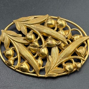 Pode incluir: Um broche oval de metal em tom dourado com um design em relevo detalhado de flores de lírio-do-vale e folhas alongadas. A peça tem um acabamento polido com texturas intrincadas na folhagem e flores arredondadas em forma de sino.