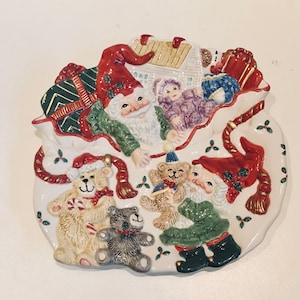 May include: A white ceramic Christmas plate with a Santa Claus and two elves carrying teddy bears and gifts. The plate is decorated with holly and gold trim.