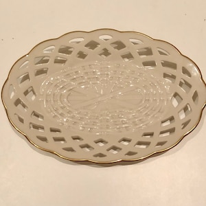 Può includere: Un piatto ovale in ceramica bianca con un bordo dorato e un motivo intrecciato. Il piatto ha un centro rialzato con un motivo intrecciato.