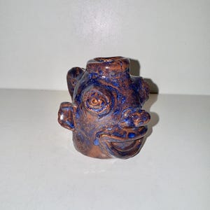 May include: A brown and blue ceramic vase shaped like a face with a wide smile and one large eye.