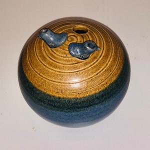 Puede incluir: Un jarrón de cerámica esférico, con tonos marrones y azules. Dos pequeñas decoraciones azules con forma de pájaro adornan la parte superior. El jarrón tiene un diseño circular y un agujero central. Diseño de cerámica.