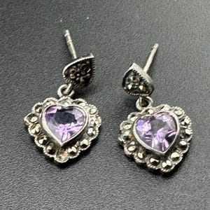 Puede incluir: Un par de pendientes colgantes plateados. Cada pendiente presenta una piedra preciosa morada en forma de corazón, rodeada por un borde decorativo. Los pendientes son de tipo de poste y tuerca.