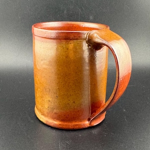 Puede incluir: Una taza de cerámica hecha a mano con un asa grande. La taza tiene forma cilíndrica y está esmaltada en tonos marrones y naranjas, con un acabado brillante. La taza es adecuada para bebidas frías o calientes.