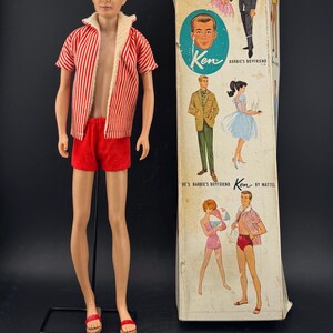 May include: Vintage Ken doll in a red and white striped shirt, red shorts, and sandals. The doll stands beside its original box, illustrated with Ken and Barbie images. The box is labelled "Ken, Barbie's Boyfriend".